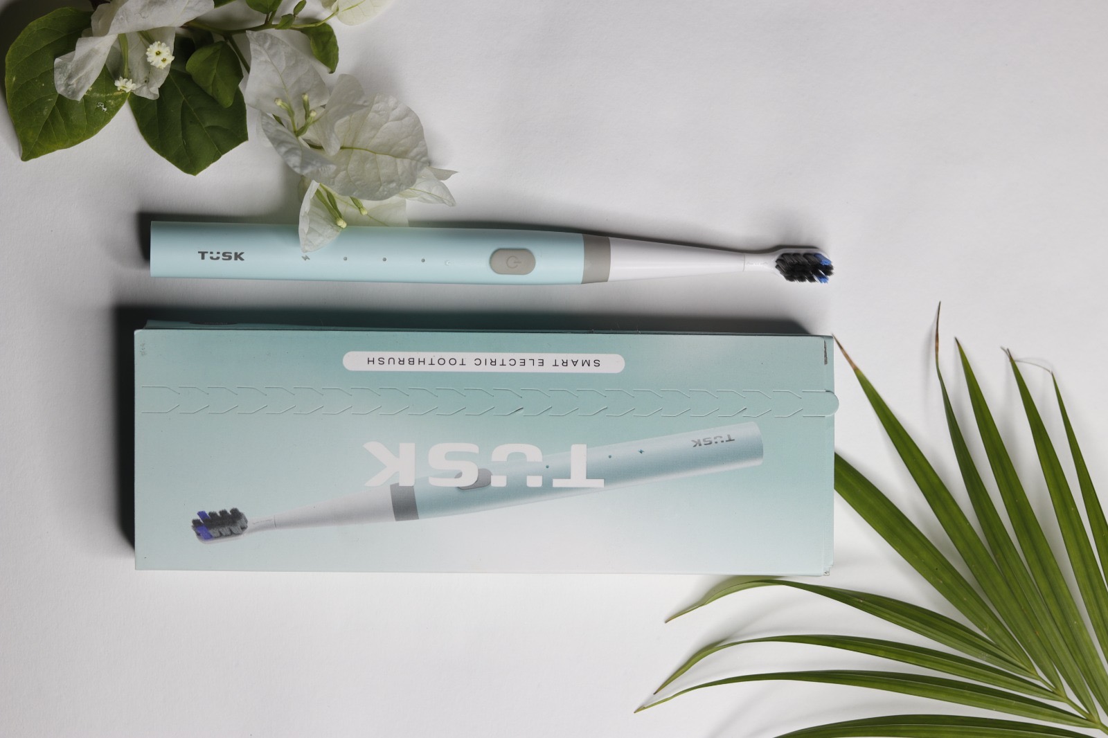 TUSK toothbrush and packaging styled with white flowers and a palm leaf on a white surface