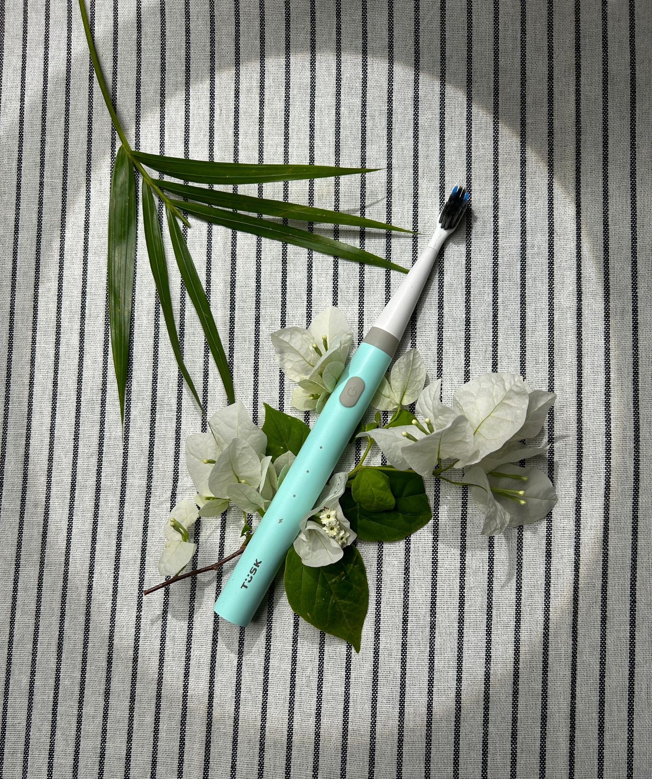 TUSK toothbrush styled with white flowers and a palm leaf on striped fabric, a lifestyle test crop